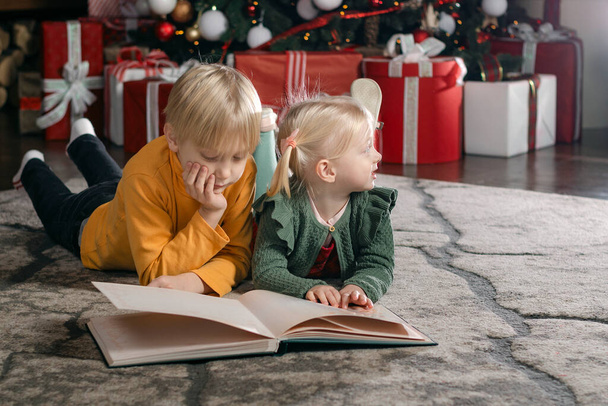Blond boy and sister with book near the Christmas tree and gifts. Siblings children on Christmas Eve - Zdjęcie, obraz
