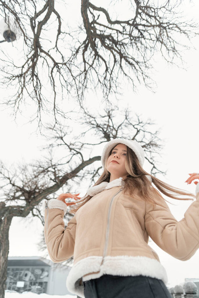 Long-haired girl in trendy white panama and short beige sheepskin coat walks along alley of park and enjoys winter day. - Fotó, kép