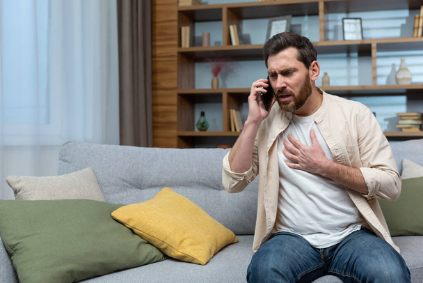 Hombre teniendo un ataque al corazón en casa, paciente llamando al médico usando el teléfono, sentado en el sofá en la sala de estar en casa. - Foto, Imagen