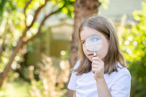 Curious clever elementary school age child, girl looking at the camera through a magnifying glass, holding a loupe in hand, one eye closed, one visible, copy space, blurred background. Young explorer - Photo, Image