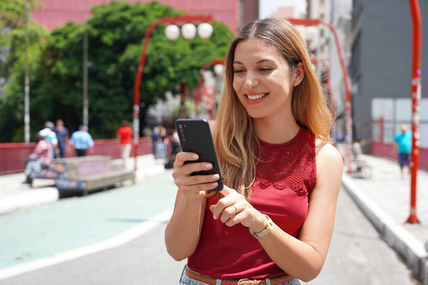 Retrato de una brasileña sonriente caminando en Sao Paulo City escribiendo en un smartphone con fondo borroso - Foto, Imagen