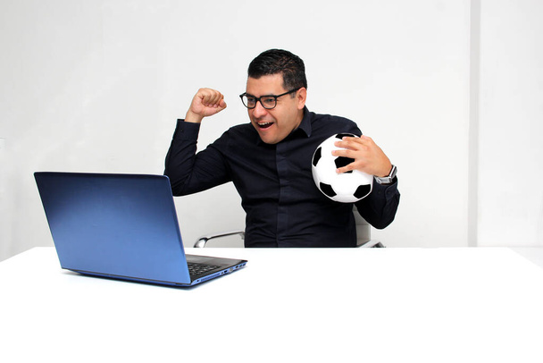 Latino volwassen kantoor man kijkt voetbal wedstrijden op zijn werk laptop tijdens kantooruren in de ochtend, hij ziet hem opgewonden, nerveus, verrast naast zijn voetbal - Foto, afbeelding