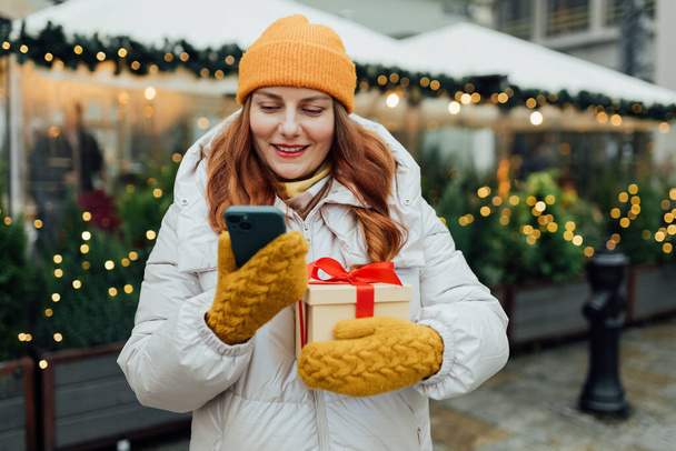 Glückliche junge schöne Frau hält eine Schachtel Geschenk und sendet SMS vom Telefon in der Nähe Weihnachtsdekoration - Foto, Bild