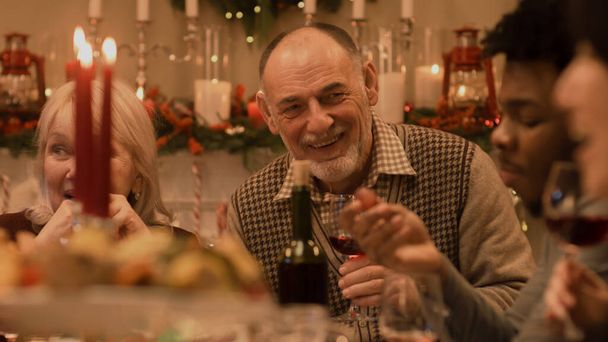Szczęśliwa rodzina świętuje Boże Narodzenie lub Nowy Rok 2023, rozmawiając i jedząc. Starsi dziadkowie się uśmiechają. Obsługiwany stół świąteczny z naczyniami i świecami. Ciepła atmosfera rodzinnej świątecznej kolacji w domu. - Zdjęcie, obraz