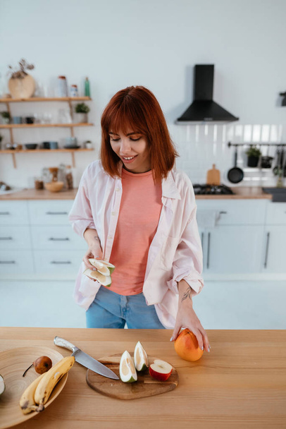 Mulher saudável feliz corte de frutas em uma tábua de madeira ao fazer café da manhã em uma cozinha
 - Foto, Imagem
