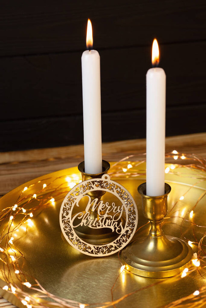 Свечи в бронзовых подсвечниках и деревянная тарелка с рождественскими надписями на золотой подставке. Черный деревянный фон. Свечи и световые фонари - Фото, изображение