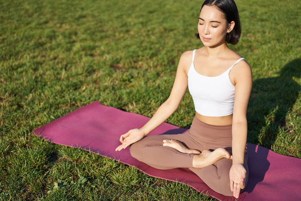 Portrait of smiling asian woman meditating, doing yoga on fresh air, relaxing on rubber mat, exercising in park, breathing air, being calm. - Valokuva, kuva