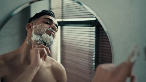 Primo piano riflesso maschile nello specchio ispanico latino indiano barbuto uomo bruna brutale ragazzo maschio in bagno spalma schiuma bianca sul viso che si prepara a radersi con gel da barba cosmetici maschili cura della pelle del viso - Filmati, video