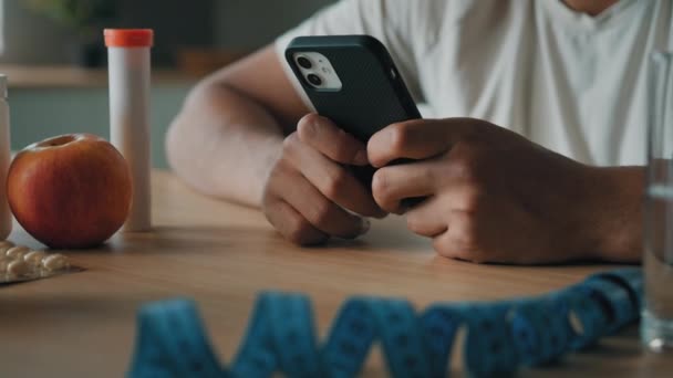 Κοντινό πλάνο αρσενικά χέρια περιήγηση στο κινητό τηλέφωνο τύπος διατροφολόγος αγνώριστος άνθρωπος αθλητικός εκπαιδευτής καταμέτρηση θερμίδων με εφαρμογή smartphone υγιεινή διατροφή fitness διατροφή με μήλο βιταμίνες χάπια μέτρο ταινία - Πλάνα, βίντεο