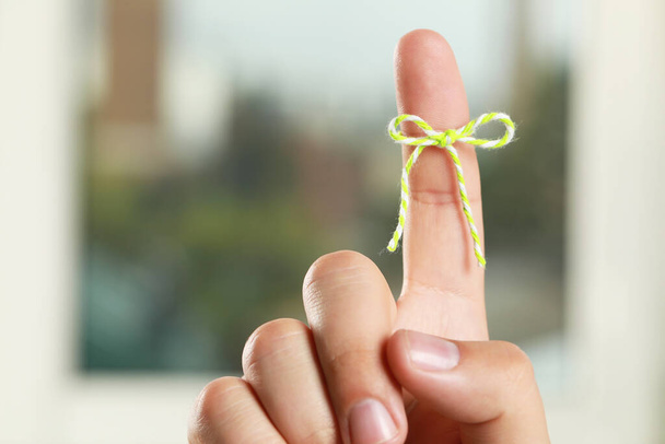 Man showing index finger with tied bow as reminder near window on blurred background, closeup - Foto, afbeelding