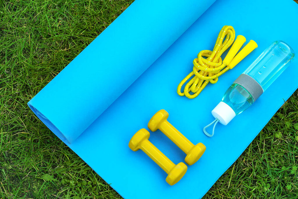 Skipping rope, dumbbells, bottle of water and fitness mat on green grass, flat lay. Morning exercise - Fotografie, Obrázek