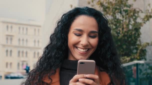 Hymyilevä onnellinen valkoihoinen latino tyttö liikenainen nainen nainen kävellä nainen kävelee kaupungin kadulla matkapuhelin älypuhelin selailu vieritys chattailuun verkossa vastaamalla viestin tekstaus sosiaalisen median sovellus - Materiaali, video