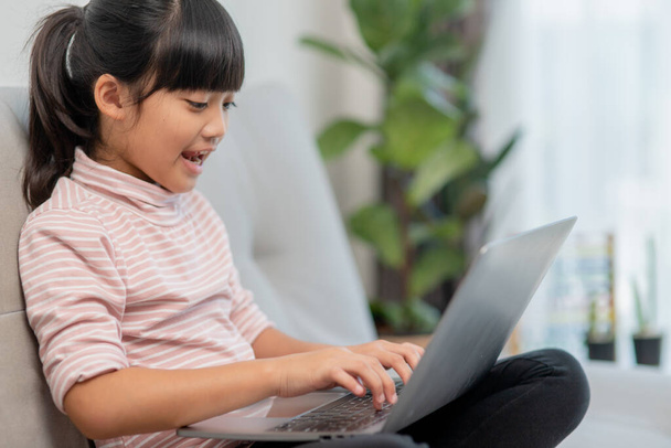 Aziatisch kleine schoolmeisje gebruik laptop computer zitten op de bank alleen thuis. Kind leren lezen online social media content, speel onderwijs lessen spel chatten met vrienden. Kinderen tech verslaving - Foto, afbeelding