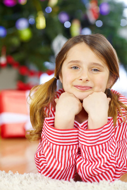 Warten in Erwartung. Ein lächelndes kleines Mädchen mit Zöpfen liegt im Schlafanzug vor dem Weihnachtsbaum - Foto, Bild