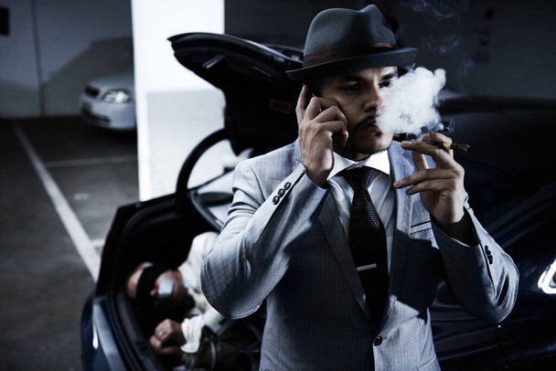 Жестокое преступление его не смущает. Высокомерный гангстер, пускающий дым в воздух, разговаривая по мобильному телефону после успешного похищения - Фото, изображение