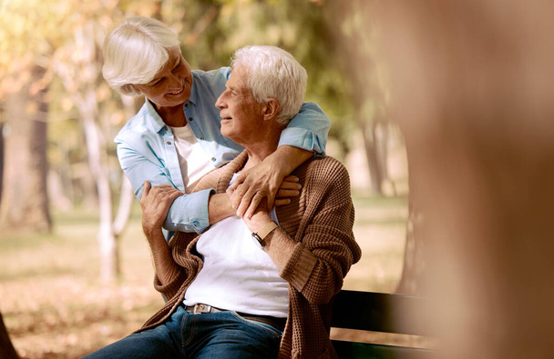 Love, senior couple and hug in park on bench, bonding and romantic together outdoor. Romance, mature man and elderly woman embrace, loving or happy for relationship, marriage and retirement in nature. - Foto, Bild