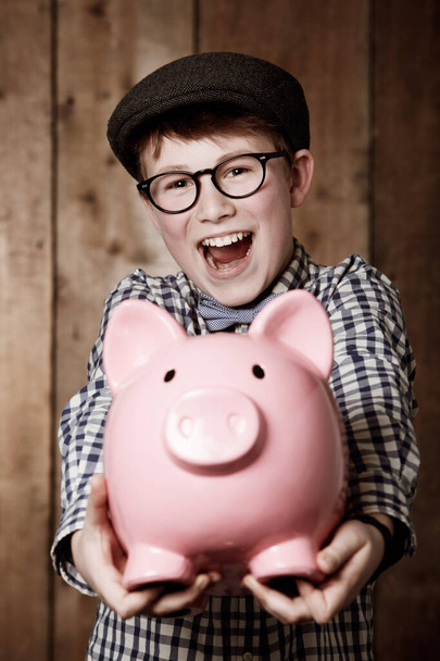 Look at all my savings. Young boy in retro clothing holding his piggybank out towards you - Foto, afbeelding