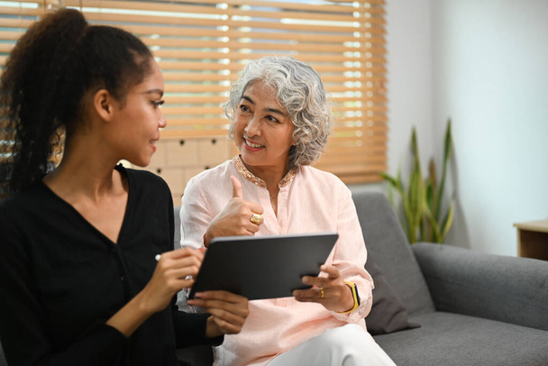 Femme âgée souriante utilisant une tablette numérique avec une fille adulte dans le salon. Personnes âgées, concept technologique. - Photo, image