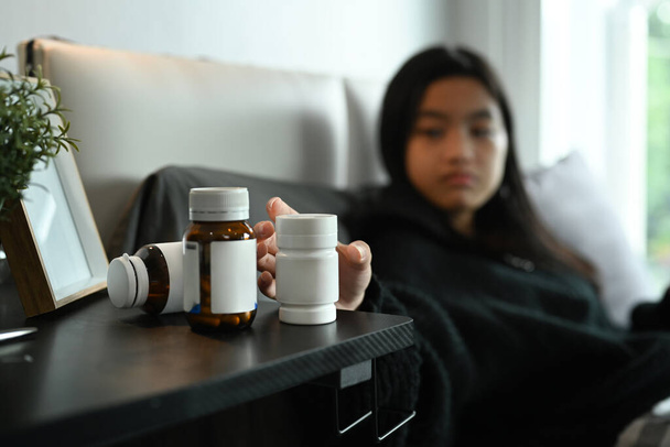 Молодая женщина принимает лекарства из прикроватной тумбочки, чтобы облегчить симптомы. Медицина, фармакология, концепция здравоохранения. - Фото, изображение