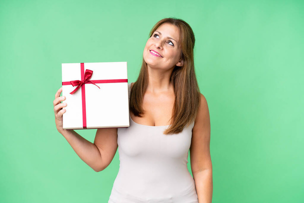 Καυκάσια γυναίκα μέσης ηλικίας που κρατά ένα δώρο πάνω από απομονωμένη φόντο κοιτάζοντας ψηλά, ενώ χαμογελά - Φωτογραφία, εικόνα