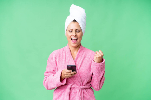 Middle age caucasian woman in a bathrobe over isolated background with phone in victory position - Foto, Imagem