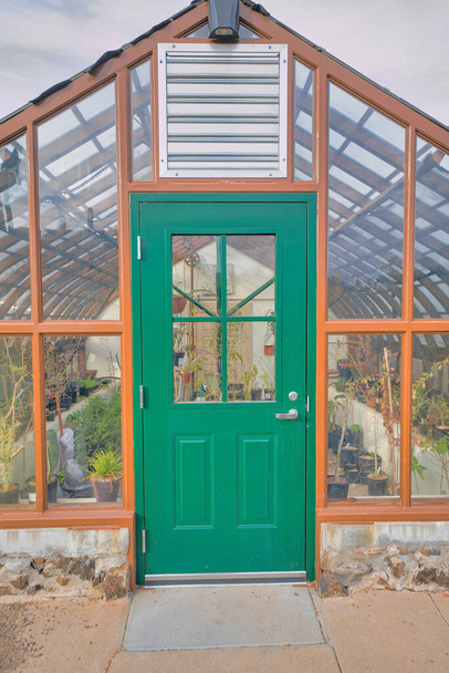 Green front door with glass panel of a greenhouse with a view of potted plants inside at Tucson, AZ. Greenhouse exterior with wood frameworks and stainless steel exhaust fan. - Photo, Image