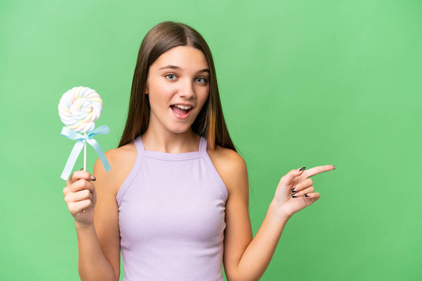 Teenager caucasian girl holding a lollipop over isolated background surprised and pointing finger to the side - Foto, imagen