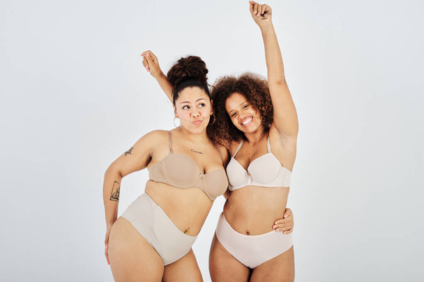 Ethnic plus size woman in lingerie putting hand on waist of undressed satisfied female model with raised arms in studio - 写真・画像