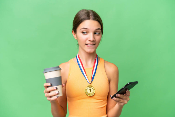 Adolescente chica caucásica con medallas sobre fondo aislado sosteniendo café para llevar y un móvil mientras piensa algo - Foto, Imagen