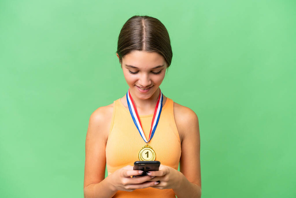 Adolescente chica caucásica con medallas sobre fondo aislado enviando un mensaje con el móvil - Foto, Imagen