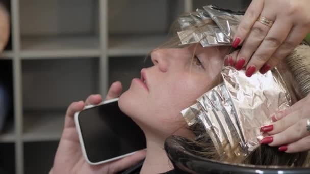 Femme occupée parlant au téléphone tout en teignant ses cheveux par un maître professionnel dans un salon de beauté avec une feuille spéciale de papier d'aluminium - Séquence, vidéo