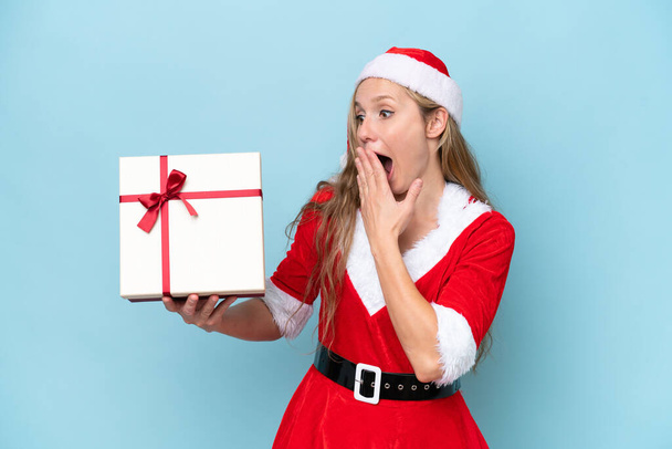 Young blonde woman dressed as Mama claus holding a present isolated on blue background with surprise and shocked facial expression - Foto, immagini
