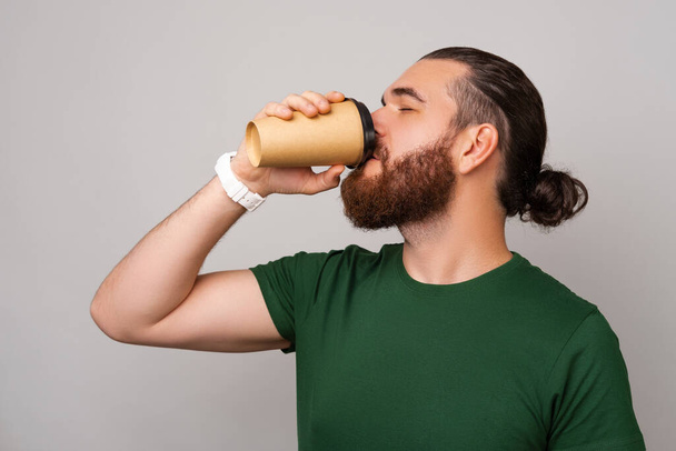 Bearded man wearing green T shirt enjoys drinking coffee from a to go cup over grey background. - Fotó, kép