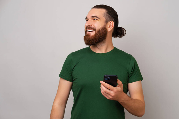 Усміхнений бородатий чоловік тримає телефон і дивиться на місце для копіювання на сірому фоні
. - Фото, зображення