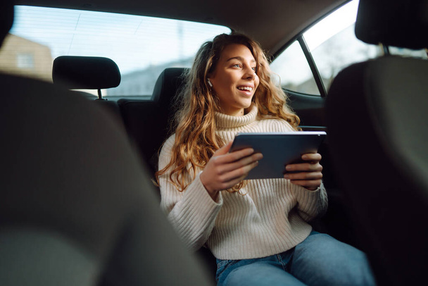 Young beautiful woman sitting in the back seat of the car with a tablet in hand. Business, technologie, internet, online concert.  - Foto, Imagen