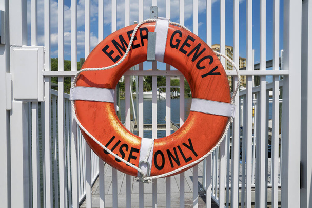 Destin, Florida Lifebuoy yüzüğü ve sadece acil durum harfleri var. Arka plandaki su manzarasına karşı beyaz bir kapının korkuluklarında can simidi asılı.. - Fotoğraf, Görsel