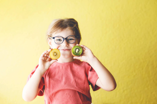 Happy little child girl with yellow and green kiwi fruits on yellow background. Preschool girl smiling. Healthy fruits for children. - Foto, immagini