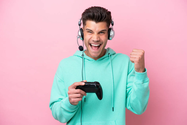 Giovane uomo caucasico bello che gioca con un controller di videogiochi isolato su sfondo rosa che celebra una vittoria nella posizione del vincitore - Foto, immagini