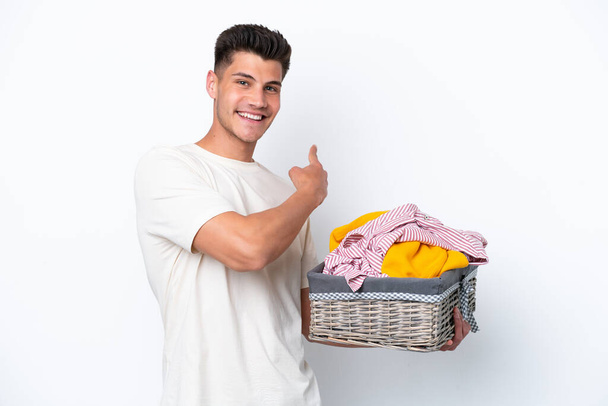 Young caucasian man holding laundry basket isolated on white background pointing back - Foto, afbeelding