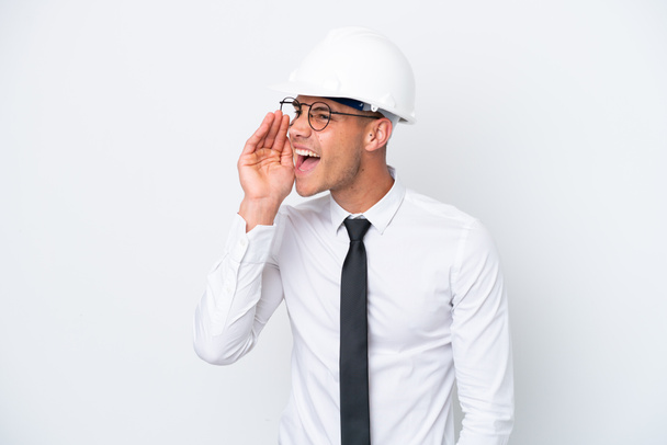 Young architect caucasian man with helmet and holding blueprints isolated on white background shouting with mouth wide open to the side - Photo, Image