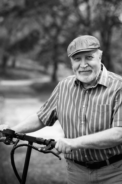 Ένας θετικός, ηλικιωμένος άντρας πήγε μια βόλτα στο πάρκο, σταμάτησε και ένα ηλεκτρικό σκούτερ στεκόταν δίπλα του. Καλοκαίρι, ηλιόλουστη μέρα. Υγιεινός τρόπος ζωής των ηλικιωμένων. Μαύρο και άσπρο  - Φωτογραφία, εικόνα