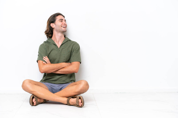 Mladý pohledný muž sedí na podlaze izolované na bílém pozadí šťastný a usmívá - Fotografie, Obrázek