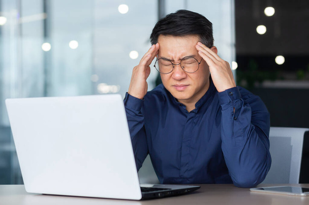 Asian businessman boss sick has severe headache, man in glasses sitting at desk using laptop at work inside office, migraine office worker investor overtired. - Photo, image