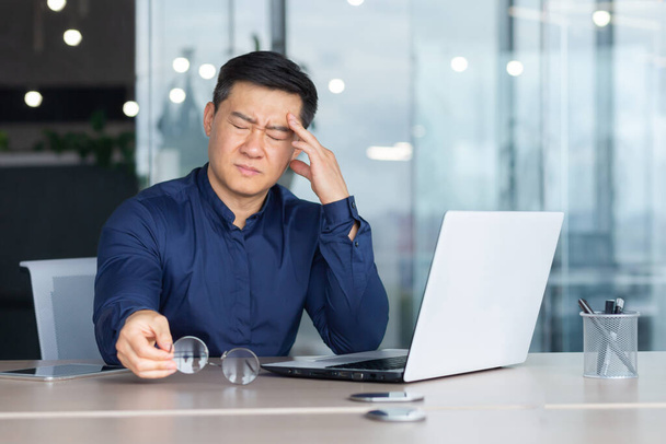 Asian businessman boss sick has severe headache, man in glasses sitting at desk using laptop at work inside office, migraine office worker investor overtired. - Foto, Bild