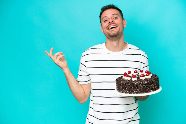 Νεαρός Καυκάσιος που κρατάει τούρτα γενεθλίων απομονωμένος σε μπλε φόντο απλώνοντας τα χέρια του στο πλάι για την πρόσκληση να έρθει - Φωτογραφία, εικόνα