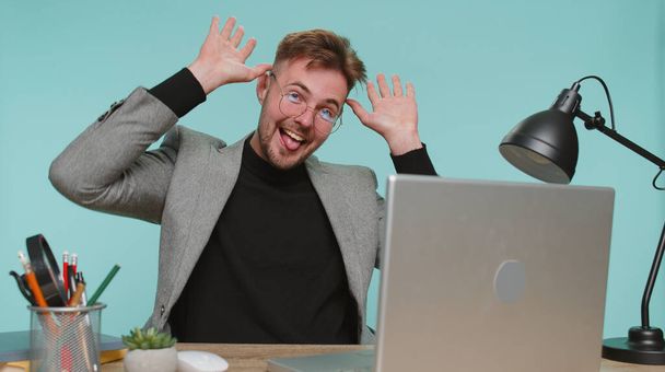 Cheerful funny bearded business man sitting at office desk showing tongue making faces at camera, fooling around, joking, aping with silly face, teasing. Young guy boy on blue studio background - Foto, Imagem