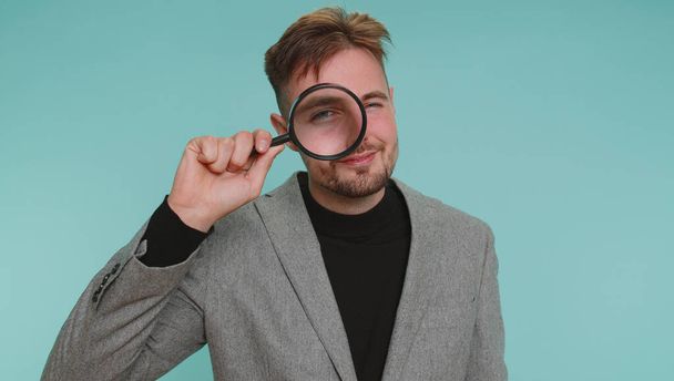 Investigator researcher scientist business man holding magnifying glass near face, looking into camera with big zoomed funny eyes, searching, analysing. Young guy isolated on blue studio background - Foto, Imagem