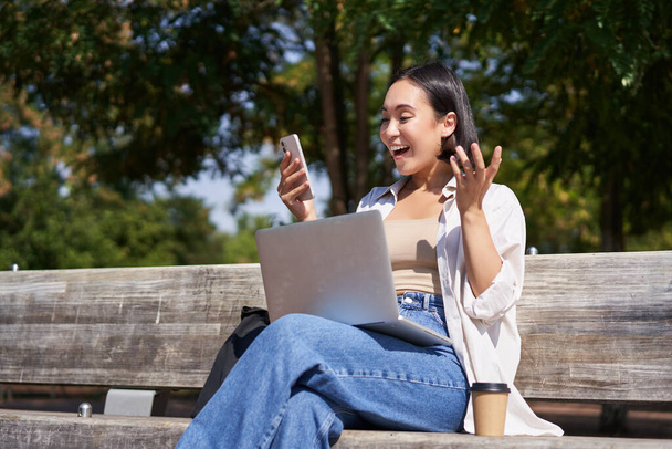 Aufgeregte junge Asiatin schaut auf ihr Smartphone, während sie mit Laptop im sonnigen Park sitzt. - Foto, Bild