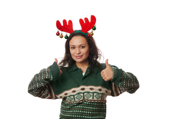 Latin American charming delightful woman, wearing a warm woolen green Xmas patterned sweater and deer antler hoop, gesturing, showing thumb up, cutely smiling looking at camera over white background - Photo, Image