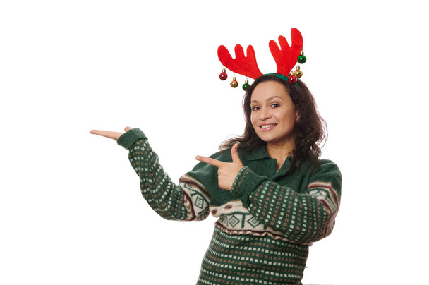 Fröhliche Brünette in weihnachtlichem grünem Pullover und Hirschgeweihkorb, die imaginären Kopierraum auf ihrer Handfläche hochhält und zeigt. Studioaufnahme auf weißem Hintergrund mit Platz für Ihren Werbetext - Foto, Bild